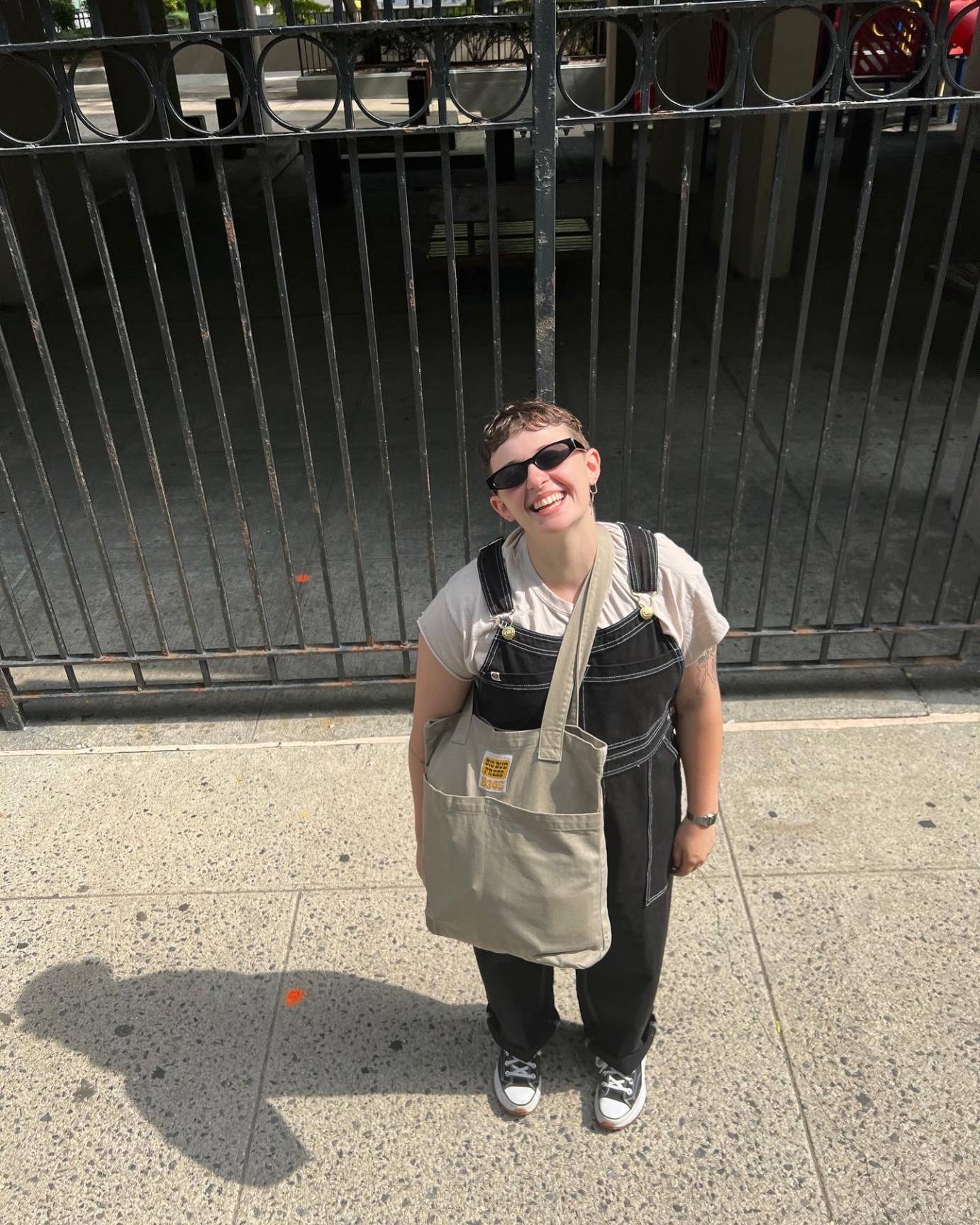 A trans woman looks up to the camera, wearing sun glasses, with a broad smile on her face. She is wearing bib overalls, a tan satchel over one shoulder and sneakers. The background is a wrought iron fence.